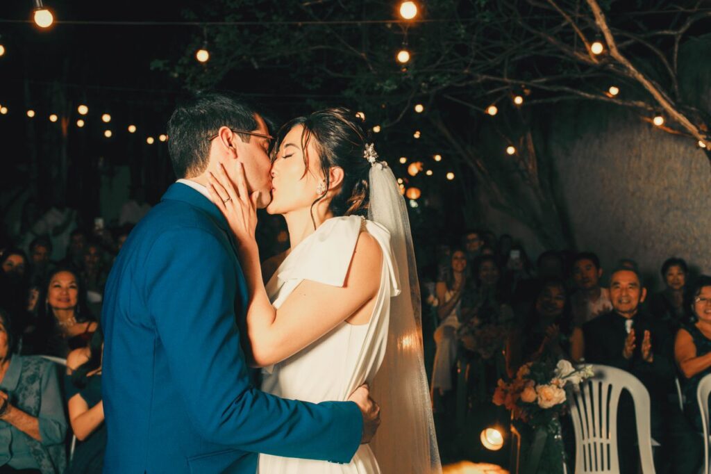 Newlywed couple sharing a romantic kiss under enchanting outdoor string lights.